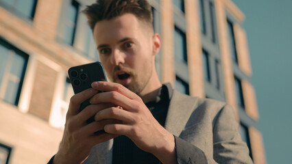 Surprised amazed Caucasian man receiving email message on phone in city outdoors shocked male guy businessman reading news on social network in Internet using smartphone on street checking SMS chat