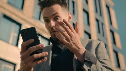 Surprised amazed Caucasian man receiving email message on phone in city outdoors shocked male guy businessman reading news on social network in Internet using smartphone on street checking SMS chat