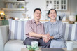 © ultramansk - A cheerful elderly couple sitting together on a cozy sofa, holding hands and smiling warmly. They enjoy their peaceful moments together, embodying love, companionship, and happiness in their home.