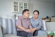© ultramansk - An elderly couple sits in their living room. The man holds his knee, appearing to be in discomfort, while his partner offers support. The image conveys care, concern, and the bond between the couple.