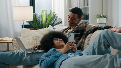 African American couple on couch man husband freelancer boyfriend working with laptop remote job with computer woman wife girlfriend scrolling mobile phone app social media at home sofa happy family