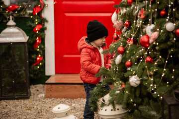 Smiling toddler boy in red jacket stands beside decorated vintage car with a wreath on the grille. A 