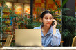 © M Alfan Setyawan - happy asian woman enjoy working on laptop and talking on mobile phone while sitting in outdoor cafe with green plant interior