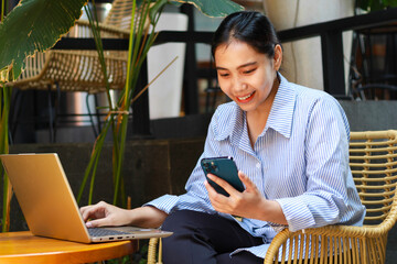 happy asian woman looking at smartphone while working remotely from cafe with laptop on wooden table