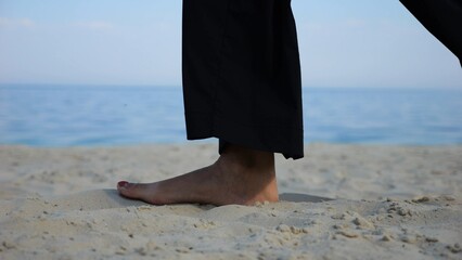 Female feet walking on sand at the beach with seascape at background. Legs of adult woman stepping along sea shore. Barefoot lady strolling near ocean coast. Concept of summer vacation. Low view