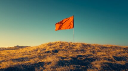 An orange flag stands alone atop a grassy mound, set against a clear blue sky, evoking a sense of solitude and tranquility in nature