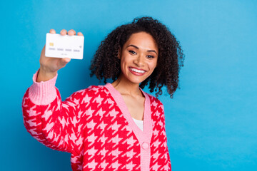 Poster - Portrait of american beautiful girl with curly hair wearing red stylish cardigan demonstrate her debit card isolated on blue color background