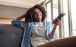 © Koegelenberg Coop/peopleimages.com - Happy, black woman and watching tv with remote on sofa for entertainment, movie or series at home. Young African, female person or streamer with smile on couch for online content or show at house