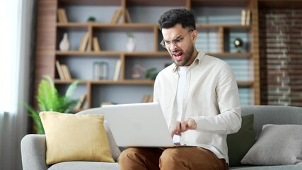 Angry frustrated young adult man having problems with software or service on laptop computer sitting on sofa in living room at home. Worried disappointed male complains about bad internet connection