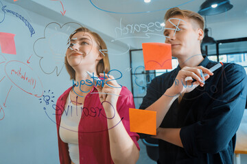 Couple of young creative start up business people brainstorming idea and solving problems by using sticky note and mind map at glass board, meeting room. discussing, working together. Immaculate.