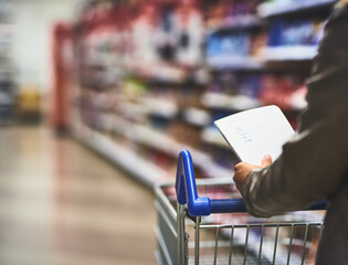 Hands, person and shopping list at food shop with wheel cart for grocery, products and prices. Closeup, customer and supermarket or convenience store for retail cost, sale and discount or promotion