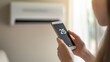 © Aziz - A person adjusts the temperature on a smartphone near an air conditioning unit.