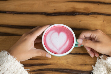 Young woman's hand holds a cup of matcha tea with raspberry or red berries, heart-shaped milk art, cappuccino coffee with red foam and heart, wooden table and decoration with plants, vegan hot drink