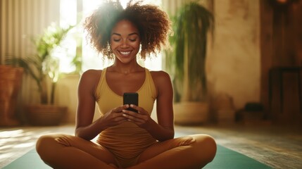 A joyful woman in yellow yoga attire sits cross-legged on a mat, smiling at her smartphone, in a sunlit room with indoor plants and warm decor elements.