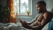© familymedia - A muscular man sitting on a bed, focused on his phone. The natural light from the window enhances his physique, adding a sense of calm and solitude to the scene.