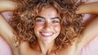 © familymedia - A smiling woman with curly hair lies relaxed on a pink surface, exuding warmth and positivity while displaying a natural, radiant aura, enhanced by her freckles.