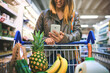 © Koegelenberg Coop/peopleimages.com - Woman, phone and aisle of supermarket for shopping, digital coupon and merchandise deal. Female person, sale and store for options on offer, retail trolley and customer check online app for price