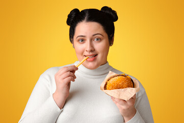 Causes Of Obesity. Young Overweight Woman Enjoying Fast Food, Eating Hamburger And French Fries, Posing Over Yellow Background With Empty Space