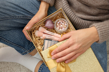 Sticker - Woman holding Christmas gift box with makeup brushes and cosmetics at home, above view