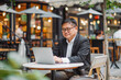 © MVProductions - Asian mid aged business man sitting by outdoor cafe table working on the laptop,concept of hard working, working outdoor in cafe, work from home, businessman, copy space