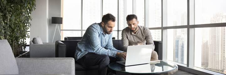 Two focused business professional men working on online project together, using laptop computer in co-working office hall, talking at laptop, discussing website presentation on Internet. Banner shot