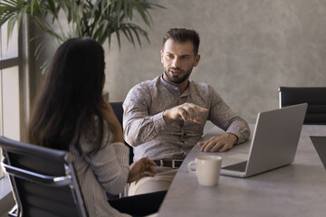 Two diverse young business professional man and woman talking at workplace, discussing work tasks, online project. Businessman speaking to female colleague at office, pointing at computer screen