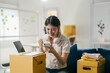 © PaeGAG - Young woman cheers happily on a couch surrounded by moving boxes, using her smartphone in a new apartment