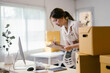 © PaeGAG - Young business owner is taking notes of new orders from her online shop. She is standing in her home office with cardboard boxes stacked behind her