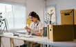 © PaeGAG - Young asian entrepreneur writing down notes while managing her business from home office, surrounded by cardboard boxes