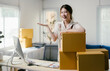 © PaeGAG - Young asian woman entrepreneur happily presents her product packaging in her cozy home office, showcasing the online business she's built