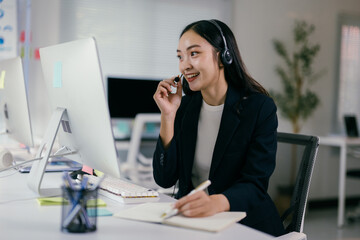 Focused asian woman in call center, wearing headset, taking notes, providing professional customer service with confidence and a smile