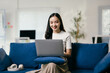 © Parichat - Young asian woman is working remotely, sitting on a blue couch in her modern apartment, looking at her laptop computer and smiling