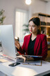 © Parichat - Focused young asian businesswoman working late in modern office, analyzing documents and typing on laptop. Red accents add color to scene