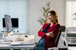 © Parichat - Young businesswoman is sitting at her desk in a modern office, smiling with her arms crossed. She is looking away from the camera, appearing confident and successful