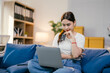 © Parichat - Asian woman happily shops online from her cozy blue couch, browsing products and services on her laptop and making secure purchases with her credit card, embodying the modern digital lifestyle