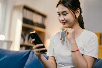 Young asian woman is sitting on her couch at home and making an online payment on her smartphone using her credit card. She is smiling because the payment process is going smoothly