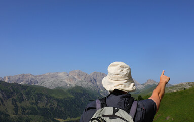 young man hiker with white hat to protect himself from the sun points to the mountains of the europe