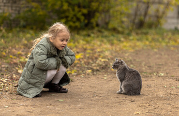 a young blond girl in a green jacket crouching on an autumn path, observing a gray cat nearby. The cat, a gray tabby with white paws, gazes at the ground with its tail curled.