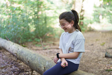 Little sad girl with glasses sits on a fallen tree in the forest. Child loneliness concept