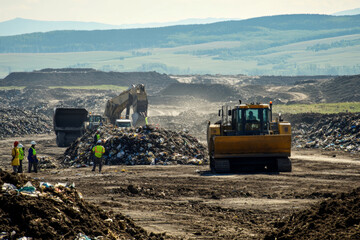 Workers in safety gear oversee heavy machinery as it moves through a large landfill, managing waste materials and maintaining the site.