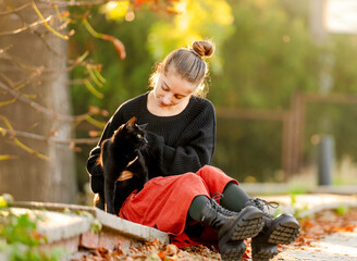 Pretty girl in red skirt hugging black cat outdoors at street with autumn leaves. Beautiful model teenager sitting with feline animal at park