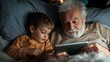 © Maximages  - A warm, intimate moment as a grandfather and his grandson share a bonding experience while browsing on a digital tablet together, enveloped in cozy blankets.