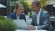 © anis - Businessman and businesswoman discussing over a laptop computer while sitting at a table outside.