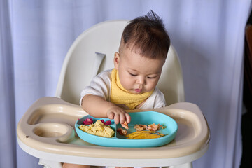 9 month old Asian baby boy is learning to eat, with food on the table, the baby eats and commands himself, it seems a bit messy