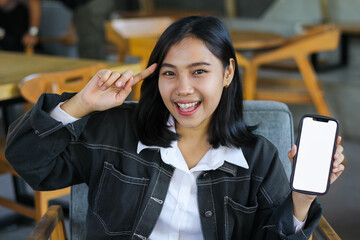 excited asian woman sitting in cafe showing smartphone empty screen and pointing finger to her head, think idea gesture