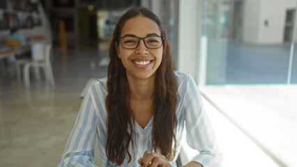 Young woman smiling confidently in an indoor office environment with glasses and long brunette hair