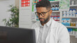 © Krakenimages.com - A focused young adult black man with glasses and a beard wearing a lab coat working in a well-organized pharmacy interior.