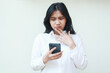 © M Alfan Setyawan - confused asian young business woman looking at mobile phone using social media and browsing internet news, worry face expression, fingers on chin wear white formal suit standing isolated background