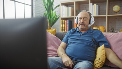 Mature man with headset sitting on couch in cozy living room, engaged in gaming or video call.