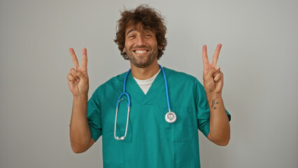 Young male doctor in green scrubs with stethoscope flashing peace signs while smiling on isolated white background